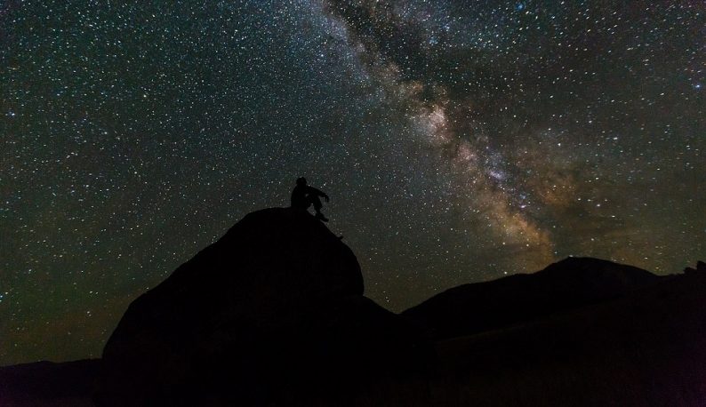 Silhueta de homem sentado em colina olhando para o céu estrelado (Foto: Banco de Imagens)