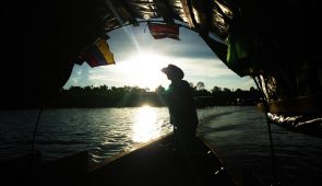 Imagem de barqueiro contra a luz do sol, passando por um túnel. No barco, estão bandeiras do Brasil, do Peru e da Colômbia (Foto: Banco de Imagens/Mauricio Goméz Quintero)