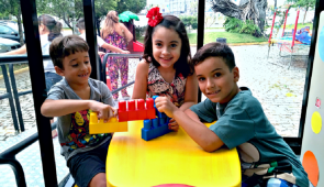 Dois meninos e uma menina sentados em uma mesa infantil amarela, brincando com peças de montar (Foto: Marília Torres)