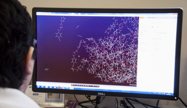 Homem usando computador, que mostra na tela uma imagem em tons roxos de moléculas químicas (Foto: Viktor Braga/UFC)