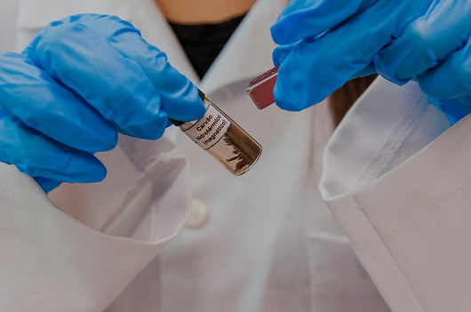 Imagem em close: pesquisadora segura um pequeno frasco contendo líquido e material carbonáceo. O carbono está sendo puxado por imã do lado exterior (Foto: David Motta/UFC)