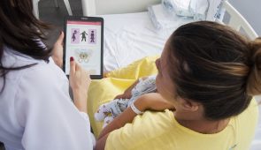 Enfermeira segura tablet e mostra aplicativo para mãe com recém-nascido nos braços (Foto: Ribamar Neto/UFC)