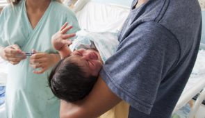Recém-nascido nos braços de um rapaz, com uma enfermeira ao fundo (Foto: Viktor Braga)