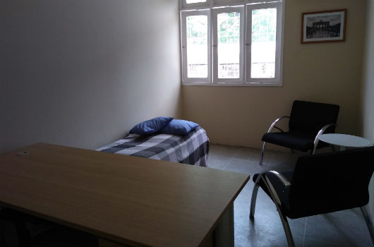 Sala com uma mesa, uma pequena cama, duas poltronas, uma janela e um quadro na parede (Foto: Luana Oliveira/PREX-UFC)