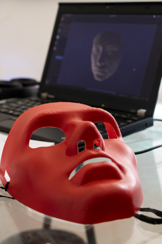 Computador ao fundo com imagem de rosto 3D e máscara em primeiro plano, reproduzindo o rosto que aparece na tela (Foto: Viktor Braga/UFC)