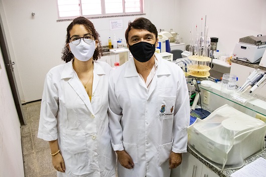 Pesquisadora Erika Luz ao lado do professor Rodrigo Vieira, ambos de jaleco, em laboratório, com diversas vidrarias e equipamentos
