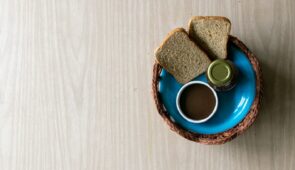 Fotografia colorida mostra vista de cima de um prato azul dentro de cesta artesanal, com um uma garrafa de vidro, pote branco com molho em tom marrom, e duas fatias de pão de forma (Foto: Viktor Braga/Agência UFC)