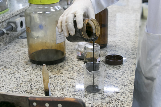 LCC, um líquido preto, sendo despejado dentro de outro recipiente por pessoa com luva branca, em uma bancada de laboratório
