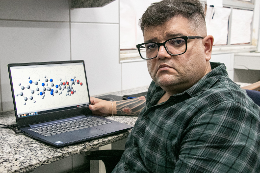 Homem branco, de óculos e camisa xadrez verde, sentado, mostra computador na qual se vê um desenho em 3D com várias bolas vermelhas e azuis conectadas, representando uma molécula no corpo humano (Foto: Ribamar Neto/UFC Informa)