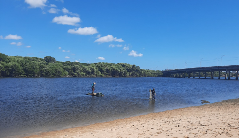 Foto mostra dois pescadores no Rio Ceará. Um deles usa chapéu de palha e está dentro um pequeno barco, enquanto o segundo está em pé, na margem, segurando uma tarrafa. Ao fundo do rio, mata verde. Do lado direito, vê-se a ponte do Rio Ceará (Foto: Equipe Lacor/Labomar/UFC)