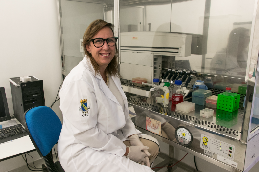 Imagem da professora Claudia Pessoa sentada, de jaleco, em laboratório