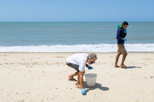 Foto: pesquisadores recolhem resíduos na praia, carregando um balde plástico