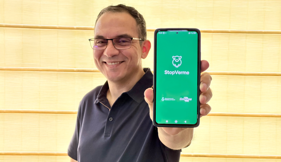 Homem de óculos e camisa polo preta posa para foto mostrando a tela de um telefone celular, onde, sobre um fundo verde, lê-se os nomes StopVerme e Embrapa e a sigla da Universidade Federal do Ceará