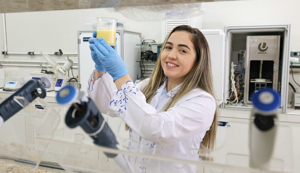 Fotografia de uma mulher sorrindo em um laboratório segurando béquer com suco de caju